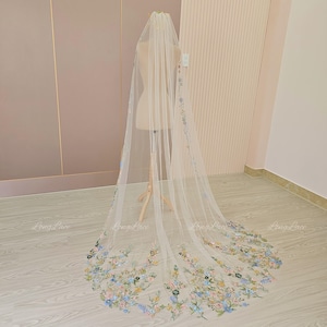 May include: A long, sheer white bridal veil with delicate floral embroidery in pastel colours. The veil is displayed on a mannequin, showcasing its length and intricate design. The floral details include blue, yellow, and pink accents.
