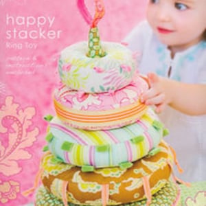 May include: A colorful ring toy with a floral and patterned design. The toy has a stack of five rings in various colors and patterns, with a small loop at the top. The packaging features the text "happy stacker Ring Toy" and "Heather Bailey Sewing Patterns".