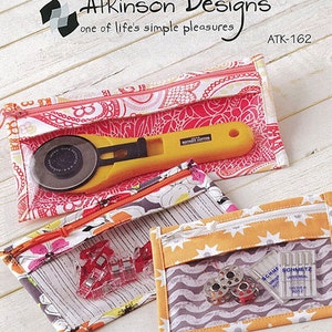 May include: Three clear-windowed fabric pouches with zippers, showcasing sewing tools. The top pouch holds a yellow rotary cutter. The middle pouch contains red clips. The bottom pouch displays bobbins and needles. The Atkinson Designs logo and text "one of life's simple pleasures" are visible.