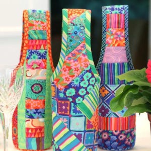 May include: Three colorful fabric wine bottle covers. Each cover features a patchwork design with vibrant floral and geometric patterns in shades of red, green, blue, and orange. The covers have a bottle-shaped design.