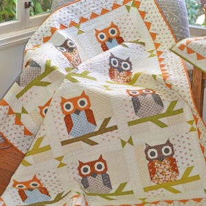 May include: A handmade quilt featuring owl designs. The quilt has a white background with orange, brown, and blue owl appliqués perched on green branches. The border is orange with a triangle pattern. The quilt is draped over a chair.
