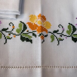 May include: White linen tablecloth with cross-stitched floral embroidery in yellow, orange, and purple. Green leaves and black stems complete the design. A stack of folded napkins is visible in the upper left corner.