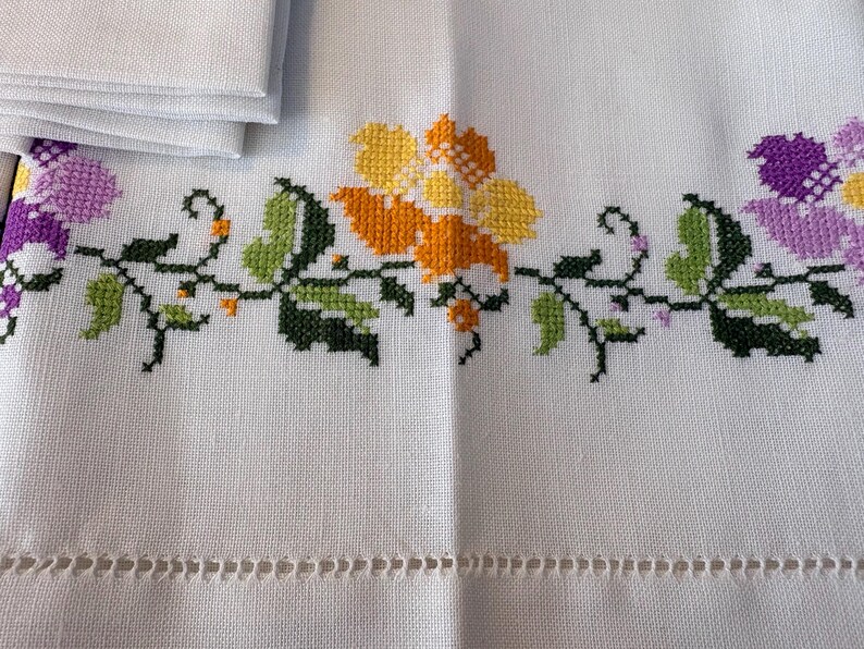 May include: White linen tablecloth with cross-stitched floral embroidery in yellow, orange, and purple. Green leaves and black stems complete the design. A stack of folded napkins is visible in the upper left corner.