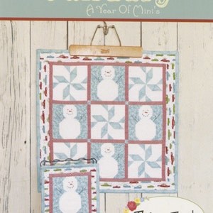 May include: A quilted wall hanging and a smaller display piece featuring snowmen and pinwheel designs. The main piece is displayed on a wooden hanger. The text "January" and "This & That" are visible.