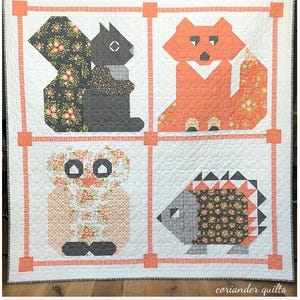 May include: A handmade quilt featuring four woodland creatures: a squirrel, fox, owl, and hedgehog. Each animal is made of geometric shapes and patterned fabric, set against a white background with a coral border. The quilt is labeled "Woodland Frolic Quilt."