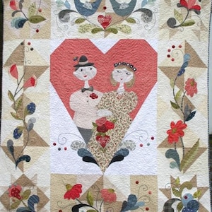 May include: A handmade quilt featuring a central heart design with a couple, surrounded by floral and bird motifs. The quilt is primarily cream, with accents of red, blue, brown, and green. The quilt is made of fabric squares and is likely for a bed or wall hanging.