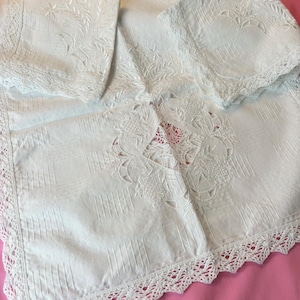 May include: White embroidered linen tablecloth and two matching napkins. The tablecloth features intricate floral and geometric designs, with a scalloped lace trim. The napkins have similar embroidered details and lace edges. The set is displayed on a pink surface.