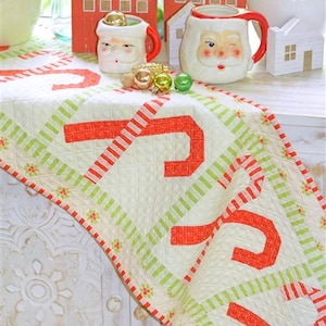 May include: A festive Christmas table runner with a candy cane design in red and white, accented with green stripes and floral details. The runner is placed on a white surface with holiday-themed decorations, including Santa mugs and wooden houses.