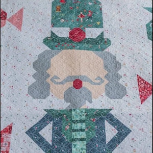 May include: A quilted Christmas-themed textile featuring a geometric design of a nutcracker. The nutcracker has a beige face, gray hair, and a red nose. The hat and jacket are patterned with green and blue fabric. The background is white with small floral accents.