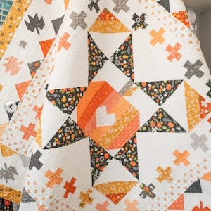 May include: A quilted blanket featuring a central star pattern composed of orange, yellow, and black floral fabric blocks. The design includes white background sections with scattered orange and gray cross shapes and leaf-shaped fabric appliqués.
