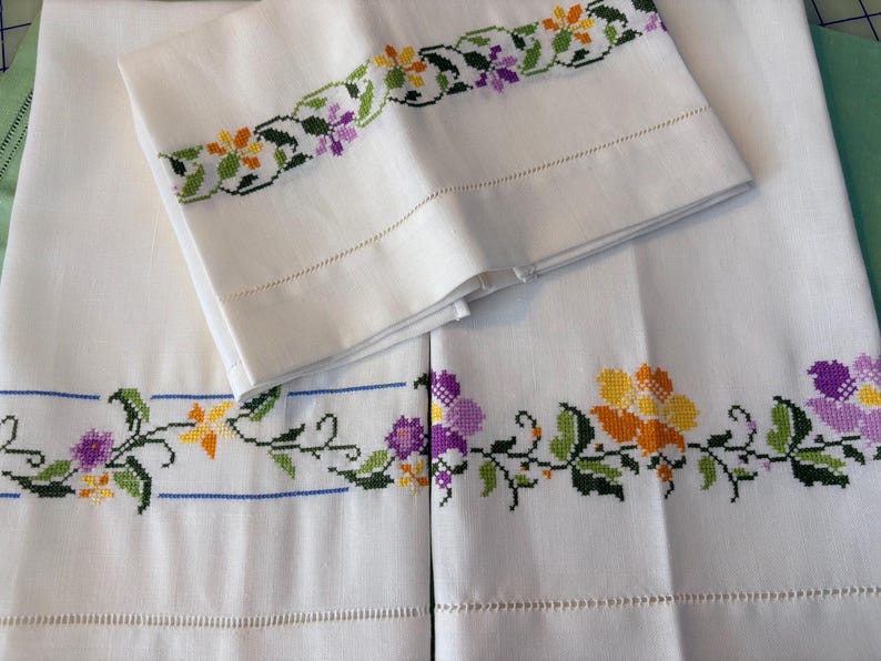 May include: White linen pillowcases and sheets with embroidered floral designs in yellow, orange, and purple. The embroidery is detailed with green leaves and stems. The pillowcases are folded on top of the sheets.