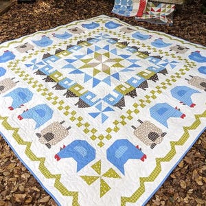 May include: A large, square quilt with a white background, featuring blue chickens and sheep, and a central starburst design in blue, green, and brown. The quilt has a geometric border and is likely handmade.