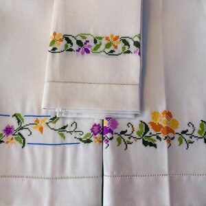 May include: White linen towels with embroidered floral borders in yellow, purple, and green. The embroidery is detailed and features a variety of flower designs. The towels are neatly folded and stacked, showcasing the craftsmanship.