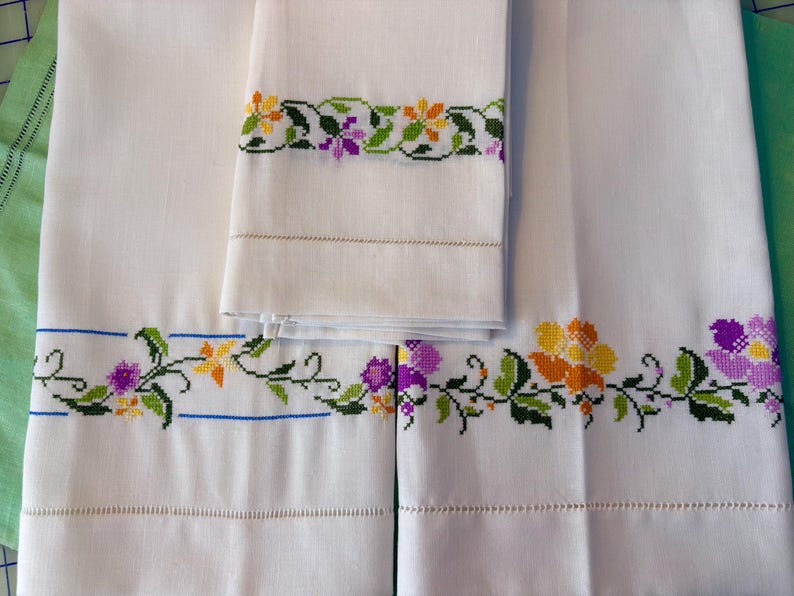 May include: White linen towels with embroidered floral borders in yellow, purple, and green. The embroidery is detailed and features a variety of flower designs. The towels are neatly folded and stacked, showcasing the craftsmanship.