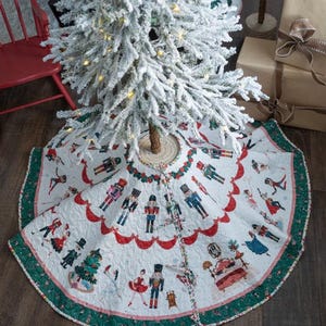 May include: A white Christmas tree skirt with a festive Nutcracker theme. The skirt features illustrations of Nutcracker soldiers and dancers, with a scalloped red border and a green trim. The tree skirt is placed around a flocked Christmas tree.