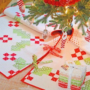 May include: A Christmas tree skirt with a red and green patchwork design. The skirt is white with red and green squares and rectangles. A stack of fabric squares is tied with a white ribbon. The tree is decorated with red ornaments and a red garland.