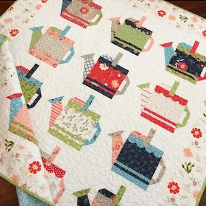 May include: A white quilt featuring a repeating pattern of colorful, patchwork teapots. The teapots are in various colors and patterns, including floral and striped designs. The quilt has a floral border and is likely handmade.