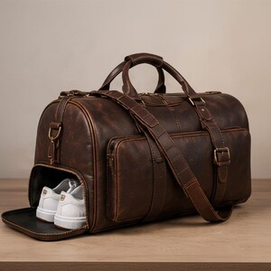 Könnte beinhalten: Eine braune Leder-Reisetasche mit separatem Schuhfach mit weißen Turnschuhen. Die Tasche hat einen oberen Griff, einen Schultergurt und Außentaschen. Das Leder hat ein Used-Finish und die Beschläge sind goldfarben.