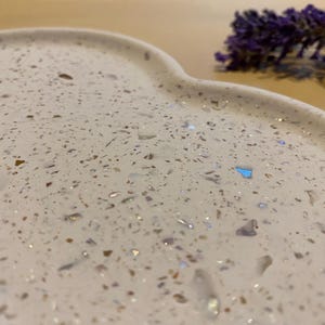 May include: A close-up of a white, speckled, heart-shaped tray with iridescent flecks. The tray has a smooth, curved edge. A sprig of purple lavender is in the background. The tray is likely made of resin or a similar material.