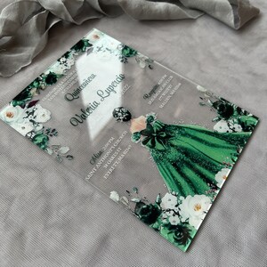 Op de afbeelding: Een transparante acryl uitnodiging voor een Quincea&ntilde;era-viering. De uitnodiging heeft een groen en wit bloemenmotief met een silhouet van een meisje in een groene jurk. De tekst op de uitnodiging luidt "Quincea&ntilde;era Valeria Lupercio 2022".