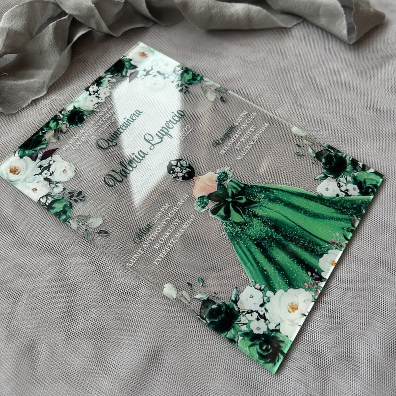 Op de afbeelding: Een transparante acryl uitnodiging voor een Quincea&ntilde;era-viering. De uitnodiging heeft een groen en wit bloemenmotief met een silhouet van een meisje in een groene jurk. De tekst op de uitnodiging luidt "Quincea&ntilde;era Valeria Lupercio 2022".