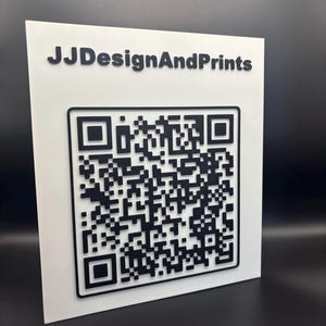 May include: A white square sign featuring a black QR code and the text "JJDesignAndPrints" in black. The sign is angled, displaying the front and side, set against a dark background.