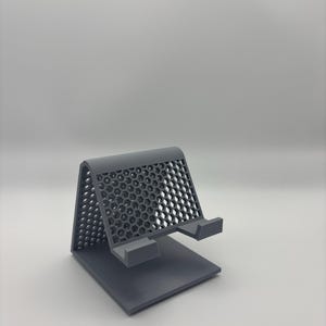 May include: A grey 3D-printed phone stand. The stand features a rectangular base, an angled back with a honeycomb pattern, and a small ledge to hold a phone. The stand is made of plastic.