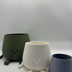 May include: Three smiling, egg-shaped planters in olive green, white, and grey. Each planter has small legs and a smiling face. The planters are suitable for small plants or succulents.