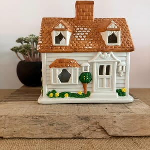 May include: A ceramic house figurine featuring white siding, a brown shingled roof with two dormer windows, a central chimney, a bay window, and a front door. The exterior is decorated with green shrubbery, small yellow flowers, and a small tree.
