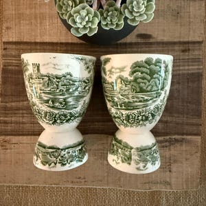 May include: Two white and green ceramic egg cups with a scenic landscape design. The cups have a pedestal base and a wide top. A decorative succulent plant in a black pot sits above the cups on a wooden surface.