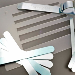 May include: A set of metal tools for creating impressions in clay or other materials. The set includes a clear plastic template with five parallel lines, a metal tool with the word "ImpressIt" printed on it, and a set of eight metal tools of varying lengths and shapes.