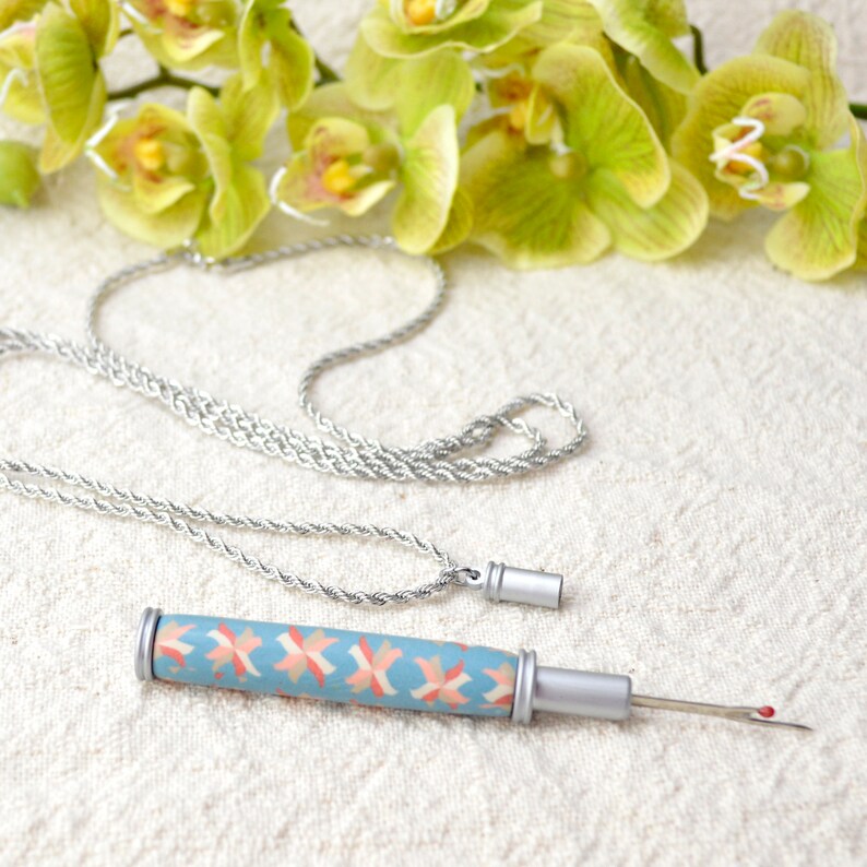 Peut inclure: Un collier argent&eacute; et un d&eacute;couseur. Le d&eacute;couseur a un manche bleu clair avec un motif g&eacute;om&eacute;trique rose et orange. La pointe de l'aiguille a une perle rouge. Des orchid&eacute;es vertes et jaunes sont en arri&egrave;re-plan.