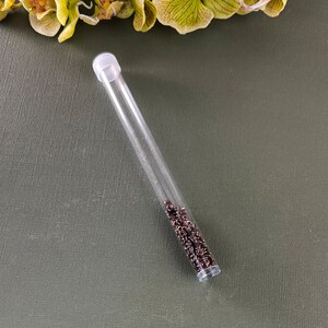 May include: A clear plastic tube filled with small, dark brown metal jump rings. The tube has a white plastic cap on one end.
