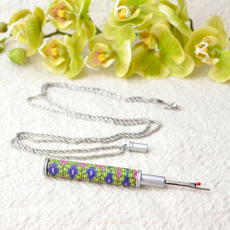 Peut inclure: Un d&eacute;couseur argent&eacute; avec un manche d&eacute;coratif orn&eacute; d'un motif g&eacute;om&eacute;trique color&eacute; vert, violet et rose. Une cha&icirc;ne argent&eacute;e est attach&eacute;e au d&eacute;couseur. Des orchid&eacute;es vertes et jaunes sont en arri&egrave;re-plan.