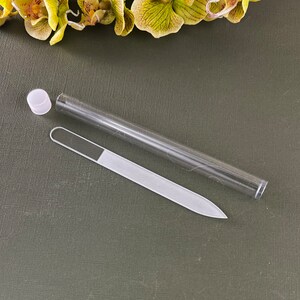 May include: A clear glass nail file with a white cap in a clear plastic tube. The file is on a green surface.