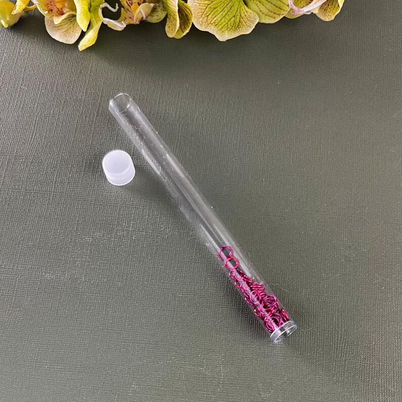 May include: A clear plastic tube with a white lid, filled with small, bright pink metal rings. The tube is lying on a dark green surface.