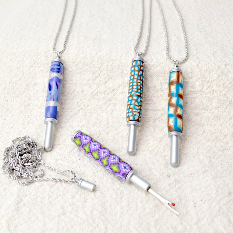 May include: Four decorative seam rippers with silver-colored metal components and colorful polymer clay handles. The handles feature various patterns in blue, purple, green, and brown. One ripper is detached, revealing the blade. Each ripper is attached to a silver chain.