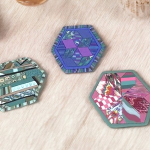 May include: Three hexagonal coasters with different geometric patterns made from fabric scraps. The coasters are green, purple, and pink.