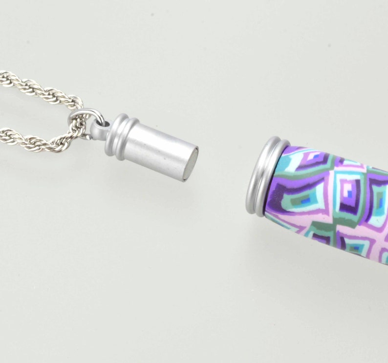 May include: A silver-colored necklace with a rope chain and a cylindrical pendant. The pendant has a geometric pattern in shades of purple, blue, and green. The pendant is separated from the chain, showing the opening.