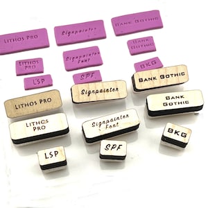 May include: A collection of wooden and rubber stamps with various fonts and styles, including "Lithos Pro", "Signpainter Font", "Bank Gothic", and "BKG".