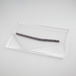 May include: A colorful, twisted polymer clay cane, featuring stripes of blue, yellow, purple, and green, rests on a clear acrylic base. The cane is a vibrant, decorative element, perfect for crafting or jewelry making.