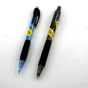 May include: Two mechanical pencils on a white surface. One pencil has a blue barrel with black accents and a yellow label that reads "TOOL". The other pencil is black with a yellow label that reads "TOOL" and "Pentel Wow! 0.7".