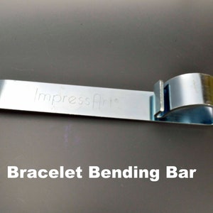 May include: A silver metal bracelet bending bar with the text "Impress Art" engraved on it. The bar is designed to help bend bracelets into shape.
