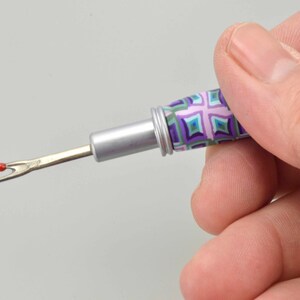 Peut inclure: Un d&eacute;couseur avec un manche &agrave; motifs dans les tons de violet, vert et bleu. La lame m&eacute;tallique a une boule rouge &agrave; l'extr&eacute;mit&eacute;. L'outil est tenu dans une main.