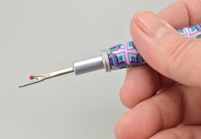 Peut inclure: Un d&eacute;couseur avec un manche &agrave; motifs dans les tons de violet, vert et bleu. La lame m&eacute;tallique a une boule rouge &agrave; l'extr&eacute;mit&eacute;. L'outil est tenu dans une main.