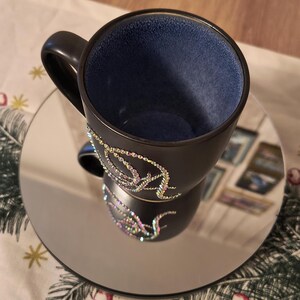 May include: A black ceramic coffee mug with a blue interior and a design of iridescent rhinestones on the side. The mug is sitting on a silver mirrored saucer.
