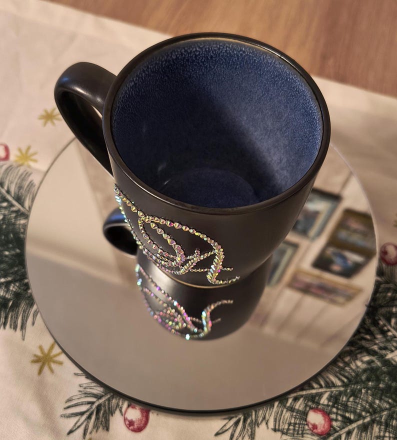 May include: A black ceramic coffee mug with a blue interior and a design of iridescent rhinestones on the side. The mug is sitting on a silver mirrored saucer.