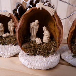 Könnte beinhalten: Ein Set aus vier Weihnachtskugeln aus Walnussschalen, die jeweils eine Weihnachtsgeschichte mit Figuren von Maria, Josef und dem Jesuskind darstellen. Die Kugeln sind mit Glitzer und Stroh verziert.