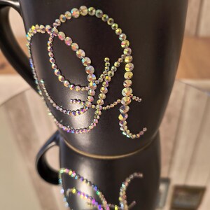 May include: A black ceramic mug with a sparkly iridescent letter "A" design on the front. The mug is stacked on top of another mug with the same design.