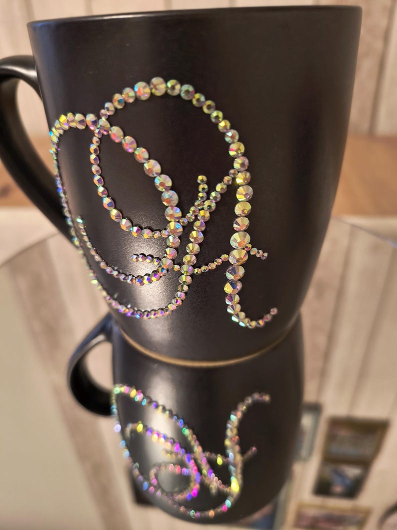 May include: A black ceramic mug with a sparkly iridescent letter "A" design on the front. The mug is stacked on top of another mug with the same design.